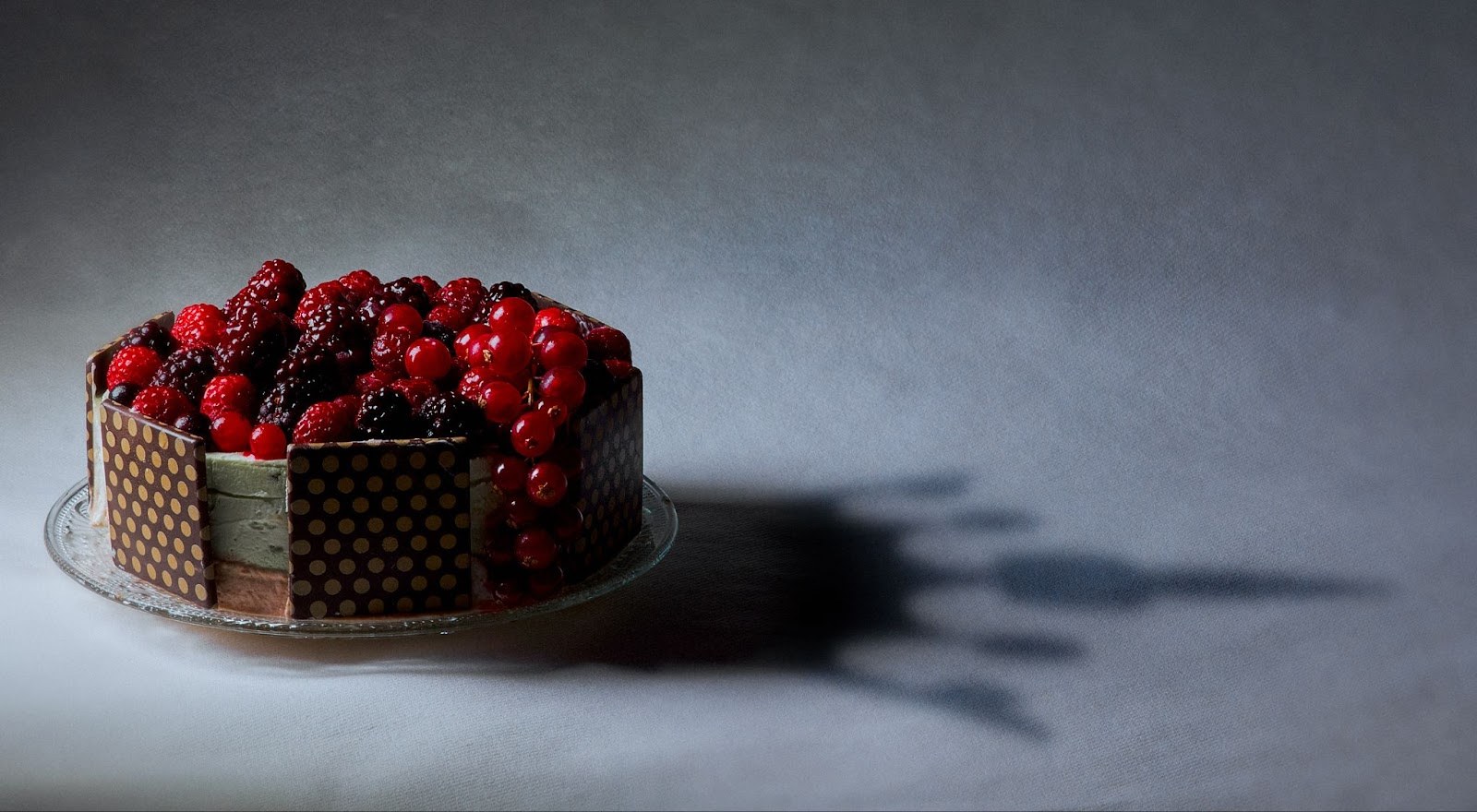 Gâteau au chocolat garni de framboises, mûres et groseilles, éclairé latéralement sur un fond gris.