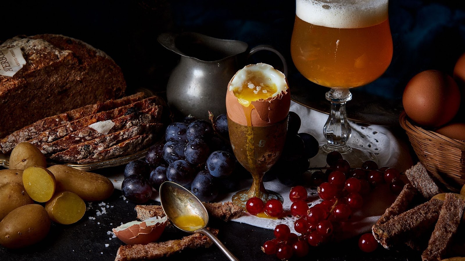 Œuf à la coque, bière ambrée, pain, pommes de terre et fruits photographiés en clair-obscur.